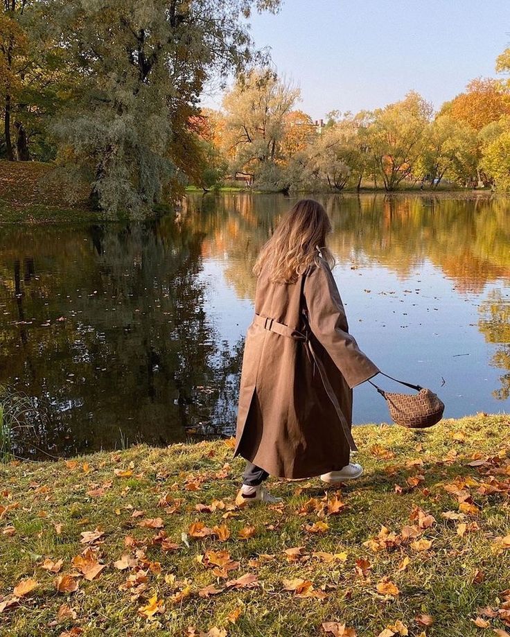 Woman in brown coat by a serene lake during autumn, with fallen leaves and reflected trees in the background.