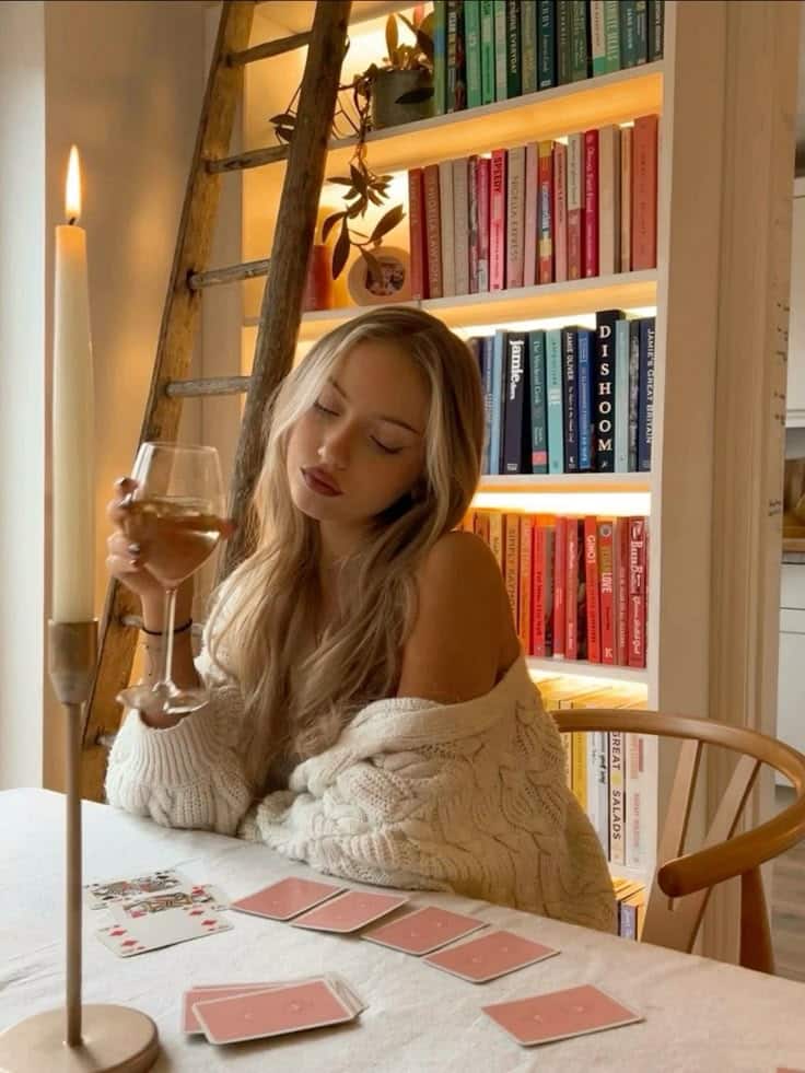 Cozy woman in a sweater sipping wine by candlelight, sitting near a bookshelf with playing cards laid out on the table.