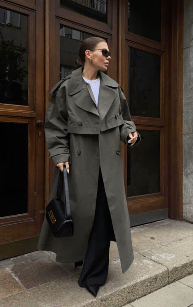 Stylish woman in a gray trench coat and sunglasses holding a black handbag on a city street.