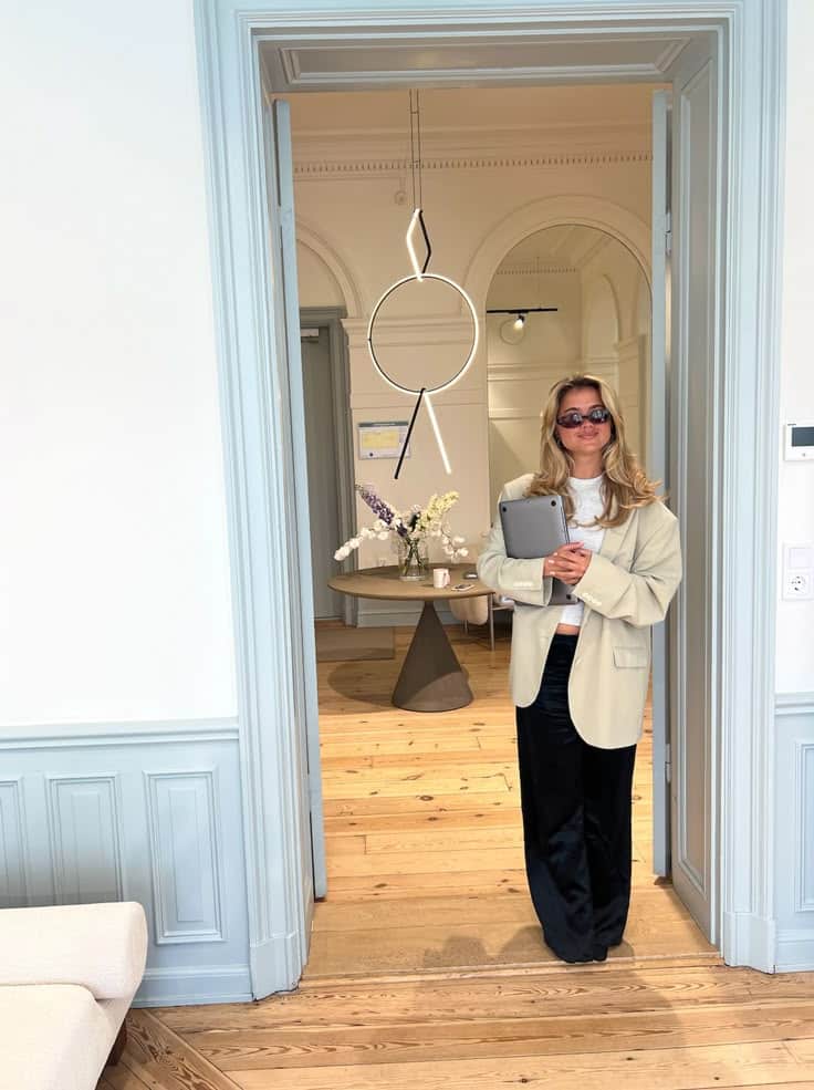 Woman in an elegant blazer holding a tablet, standing in a stylishly decorated room with modern lighting.