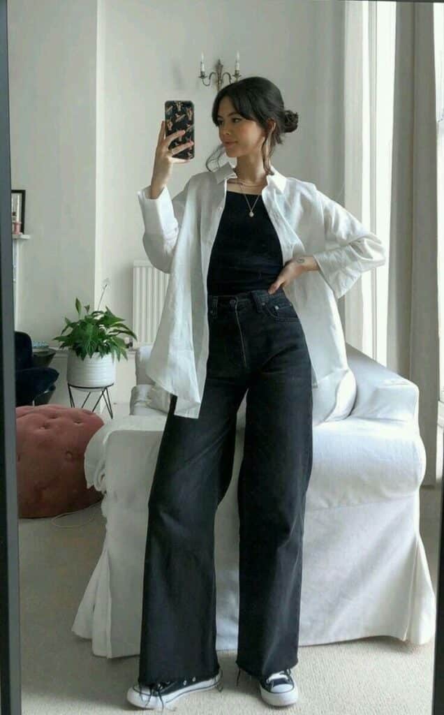 Woman in a casual outfit taking a mirror selfie, wearing a white shirt, black top, and wide-leg jeans with sneakers.
