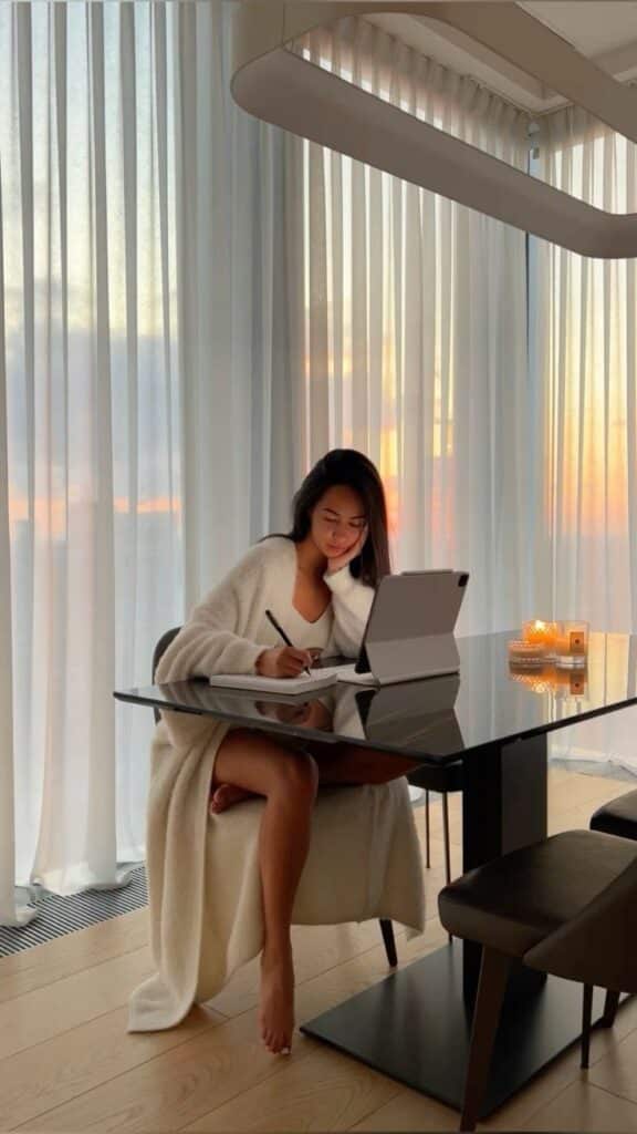 Woman writing at a modern glass table in soft evening light, cozy atmosphere with candles, tablet, and notebook.