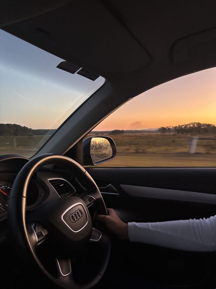 Person driving Audi during sunset, steering wheel visible, scenic countryside view.