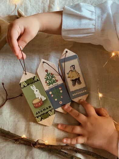 Зимняя коллекция_ Child holding vintage-style wooden ornaments on a cozy fabric background with warm fairy lights. | Sky Rye Design Child holding vintage-style wooden ornaments on a cozy fabric background with warm fairy lights.