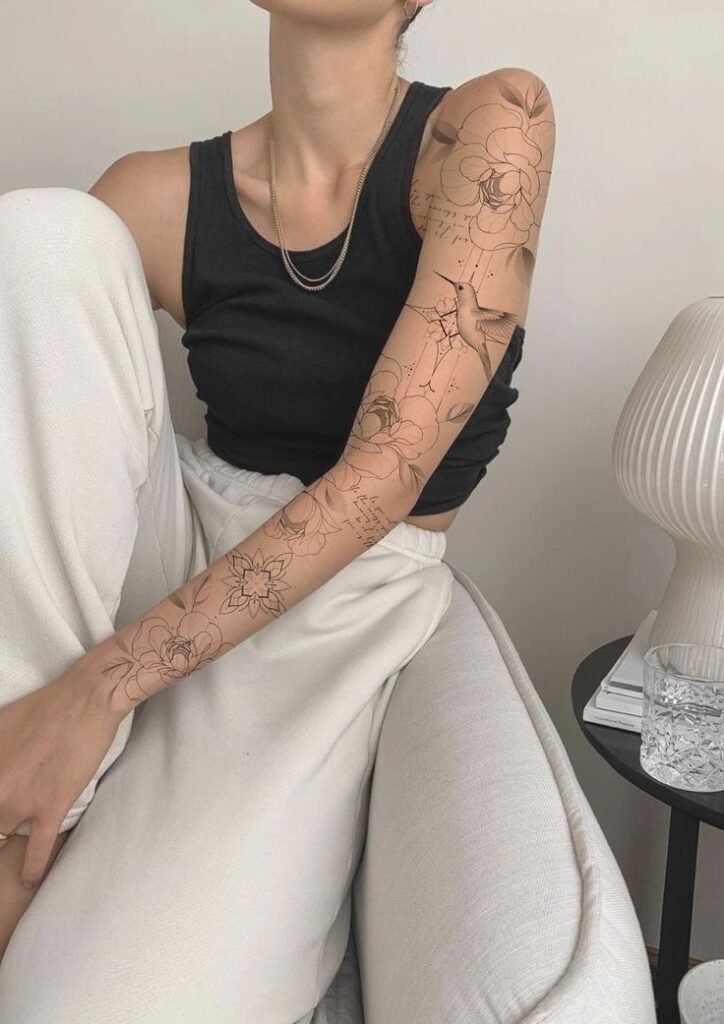 Проект женского рукава 🙌 Person with floral and hummingbird tattoo sleeve wearing a black tank top and white pants, seated casually indoors. | Sky Rye Design Person with floral and hummingbird tattoo sleeve wearing a black tank top and white pants, seated casually indoors.
