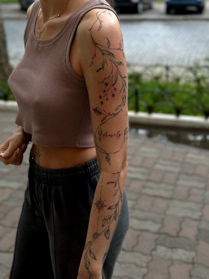 тату рукав Arm tattoo with leaves and berries design on a person wearing a sleeveless top, walking outdoors on a paved path. | Sky Rye Design Arm tattoo with leaves and berries design on a person wearing a sleeveless top, walking outdoors on a paved path.
