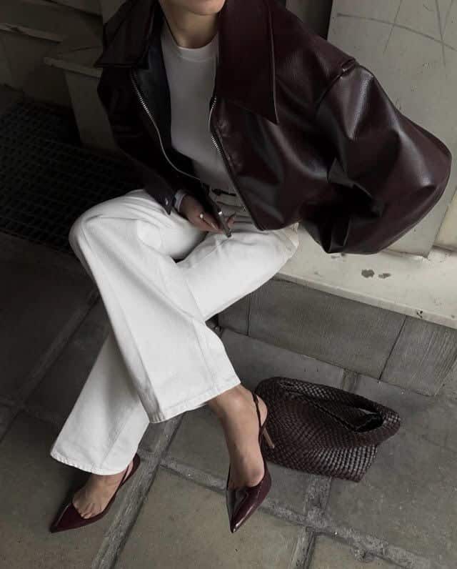 Fashionable woman in leather jacket and white pants, holding phone with woven bag and burgundy heels. Trendy street style.