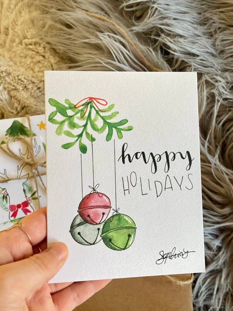 01d5179435a142d539045195ffcf7231 Hand holding a holiday card with watercolor bells and Happy Holidays message on a festive background. | Sky Rye Design Hand holding a holiday card with watercolor bells and Happy Holidays message on a festive background.
