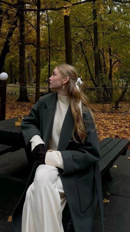 Woman in fall attire sitting on bench amidst autumn trees, wearing a dark coat and cream dress, looking to the side.
