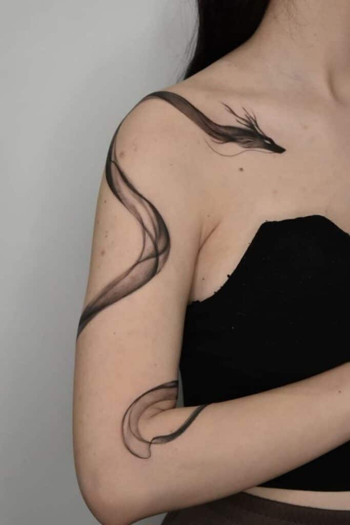 04f12b6c-c80d-4cdd-9731-df677e43c339 Tattoo of a flowing black ribbon resembling a dragon design on woman's shoulder and arm. | Sky Rye Design Tattoo of a flowing black ribbon resembling a dragon design on woman's shoulder and arm.