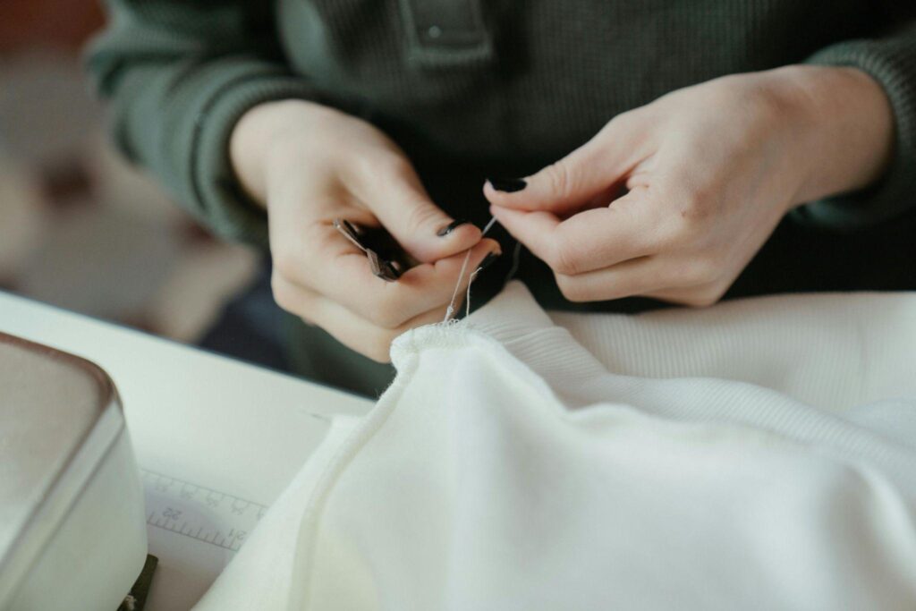 05771D1B-7D6C-4C11-AAF8-43A619A5E067 Person hand-sewing a white fabric with a needle and dark thread, focused on detailed stitching. | Sky Rye Design Person hand-sewing a white fabric with a needle and dark thread, focused on detailed stitching.