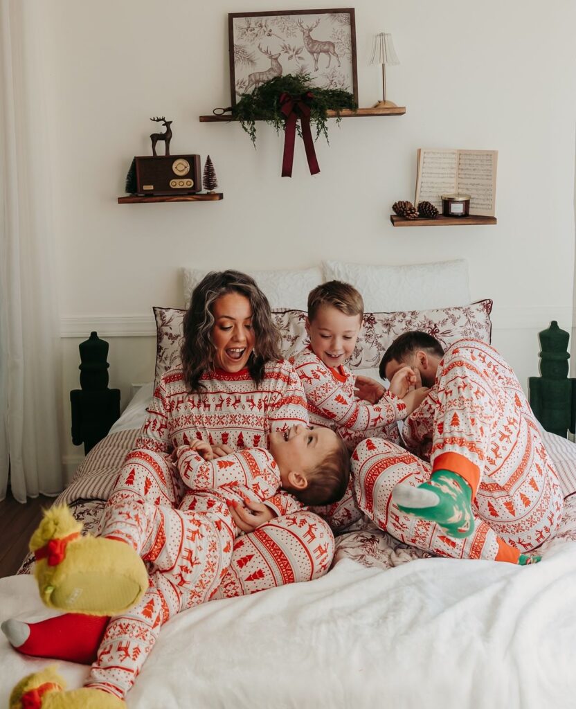 Family in matching Christmas pajamas laughing on a bed, surrounded by festive decor.