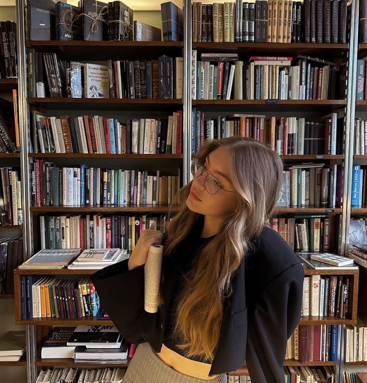 Woman with long hair and glasses in a library, holding a book and looking to the side. Cozy, intellectual setting.