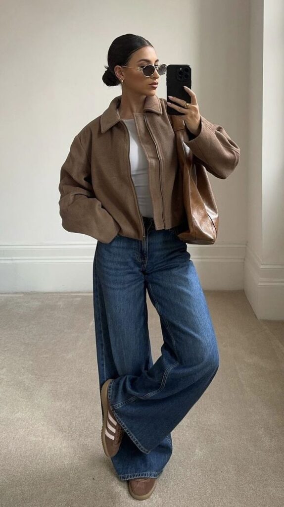 Stylish woman in sunglasses taking mirror selfie, wearing a brown jacket, wide-leg jeans, and holding a brown bag.