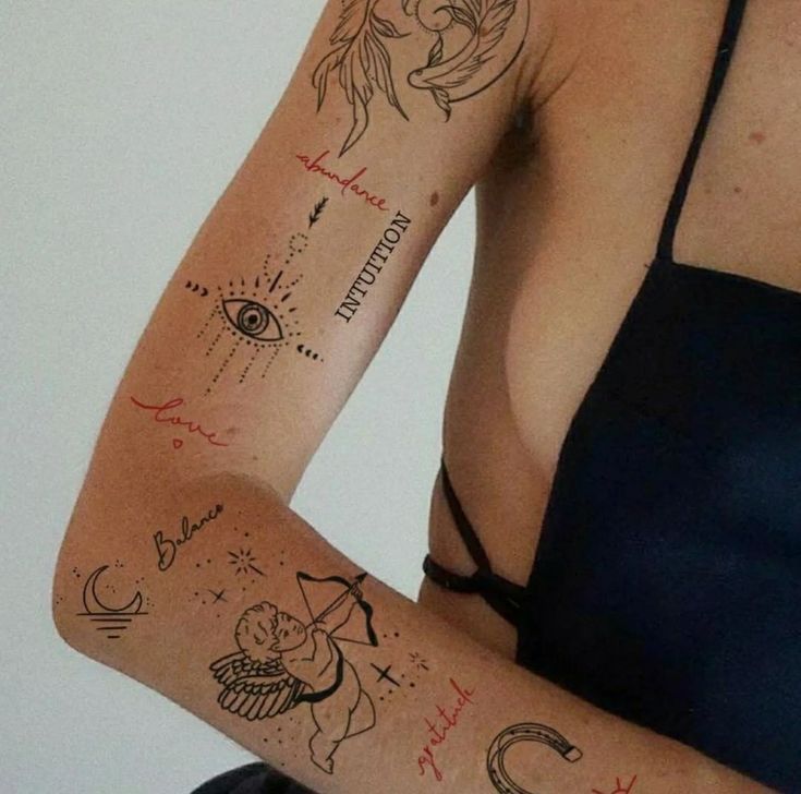 Arm adorned with tattoos: eye, moon, charms, cherub, and words like abundance, intuition, love, balance, gratitude.