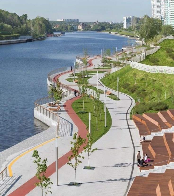 Modern riverside walkway with red path, trees, and benches, overlooking water and greenery, ideal for leisurely strolls.