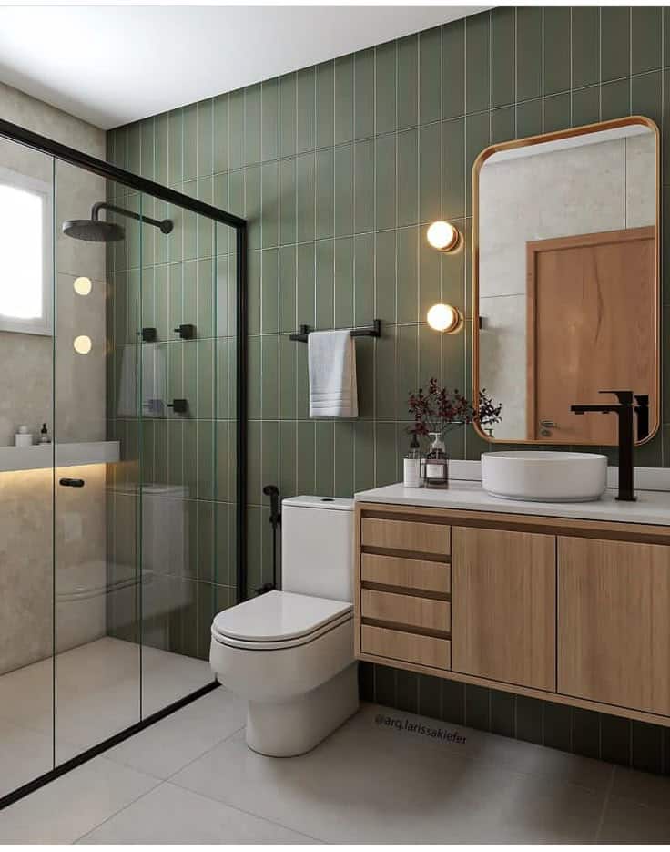 Modern bathroom with glass shower, green tiles, wooden vanity, round mirror, and decorative lighting.