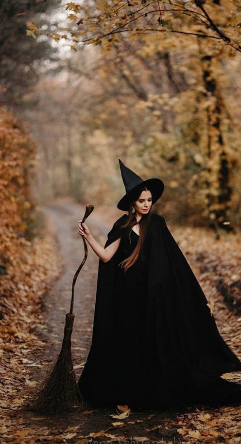 Witch in black cloak and hat holding broom, standing on a leaf-covered path in an autumn forest.
