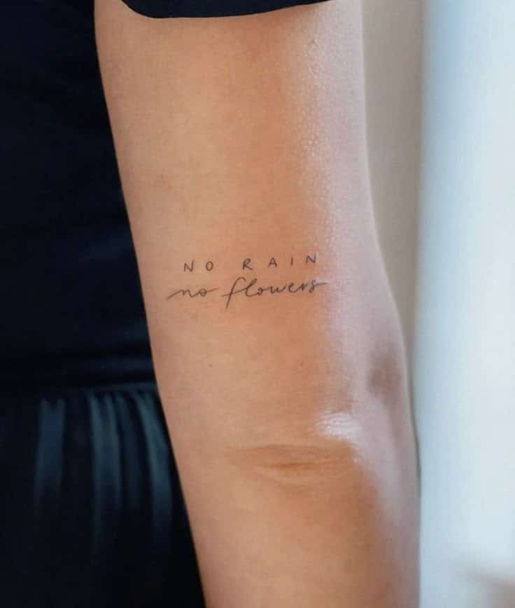 Minimalist arm tattoo reads No Rain No Flowers in cursive and uppercase, symbolizing resilience and growth.
