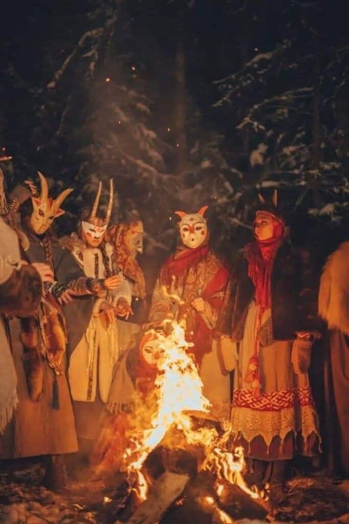 People in traditional masks gather around a campfire in a forest at night.