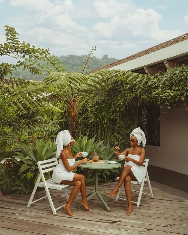 Two women in towels enjoy coffee at an outdoor patio surrounded by lush greenery.