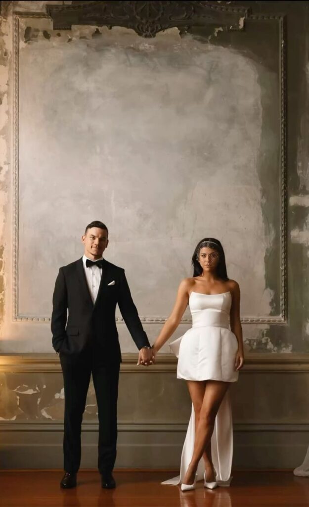 Elegant couple holding hands in formal attire, standing against a vintage textured wall.