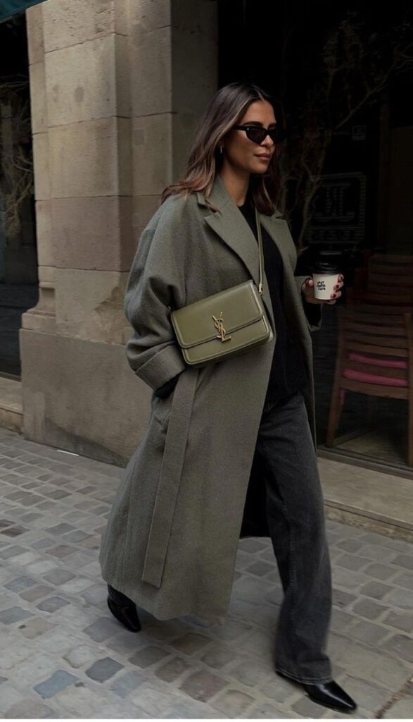 Stylish woman in sunglasses, long coat, and boots holding coffee, with a green designer bag on a city street.