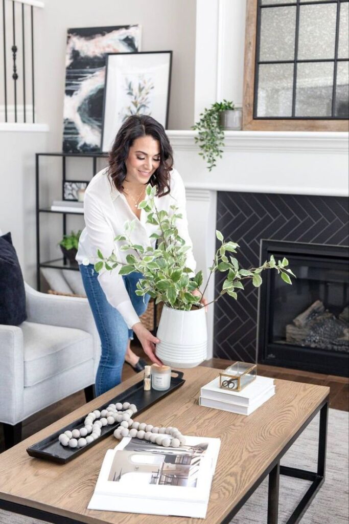 Stylish living room decor with woman placing a plant on a wooden coffee table near a cozy fireplace.
