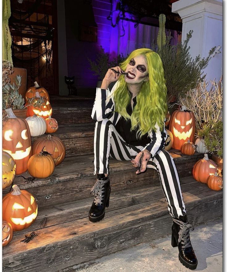 Person in green wig and striped outfit sitting by Halloween pumpkins on steps.