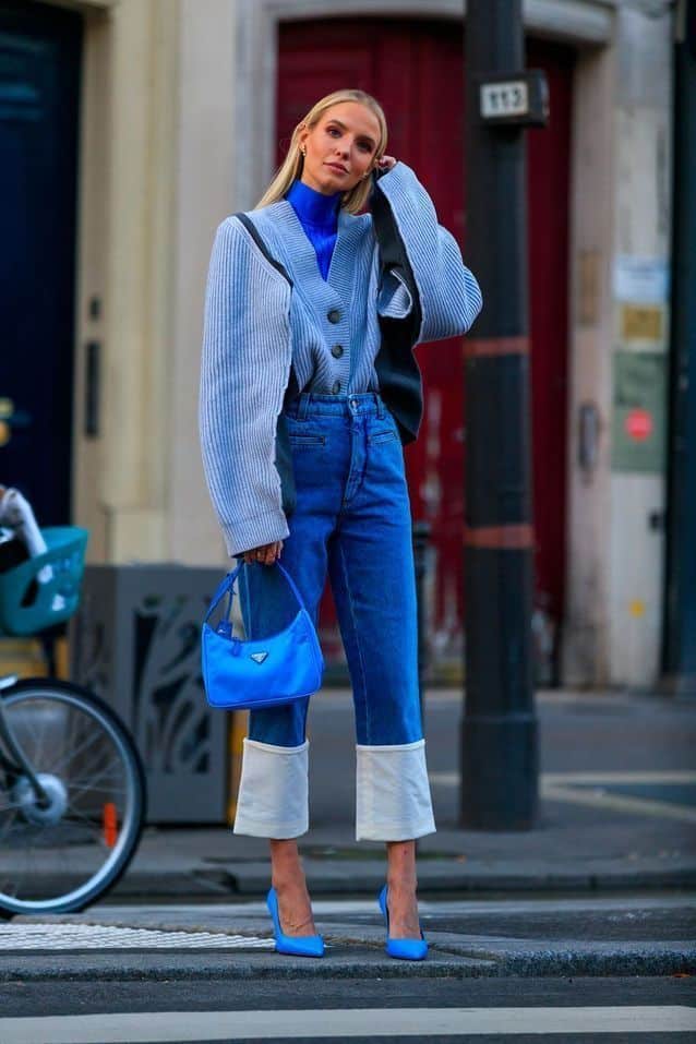Fashionable woman in blue jeans and sweater, holding a blue handbag, stands on a city street in high heels.