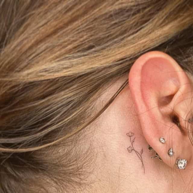 Minimalist flower tattoo behind ear with multiple earrings.