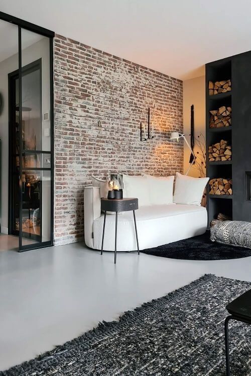 Modern living room with exposed brick wall, cozy white sofa, black side table, and stacked firewood.