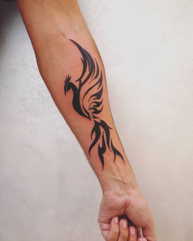 Stylish black phoenix tattoo on forearm with tribal design, symbolizing rebirth and strength against a light background.