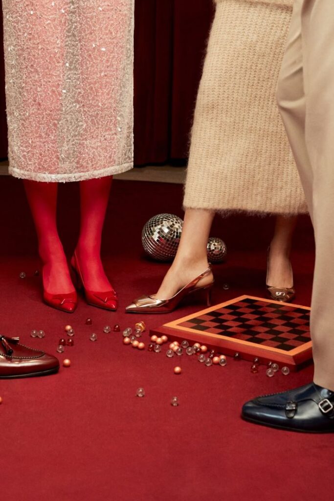 Fashionable footwear and elegant outfits surround a chessboard on a red carpet with scattered beads and disco balls.