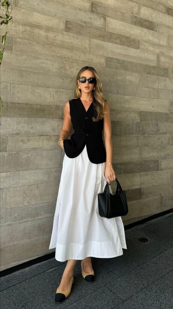 Stylish woman in black vest and white skirt holding handbag, wearing sunglasses and heels, against a stone wall.