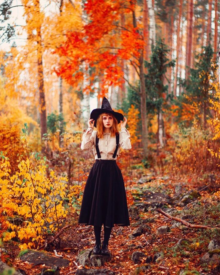 Young woman in witch costume standing in an autumn forest, surrounded by vibrant orange and yellow foliage.