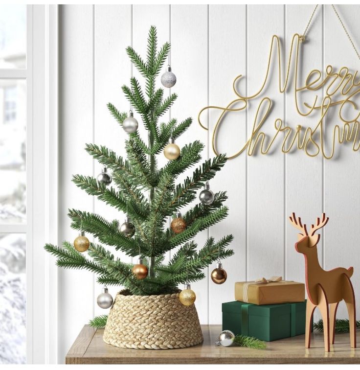 Small Christmas tree with ornaments on a table, surrounded by gifts, reindeer figurine, and Merry Christmas sign.