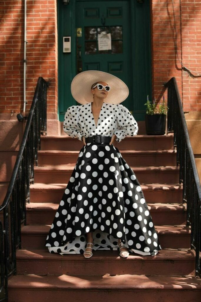 Woman in polka dot dress and wide-brim hat stands on steps. Fashionable outfit with sunglasses.
