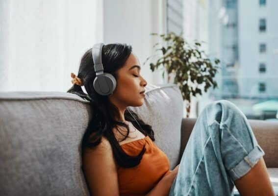 Woman relaxing on a sofa, eyes closed, wearing headphones and an orange top, enjoying music in a cozy living room.