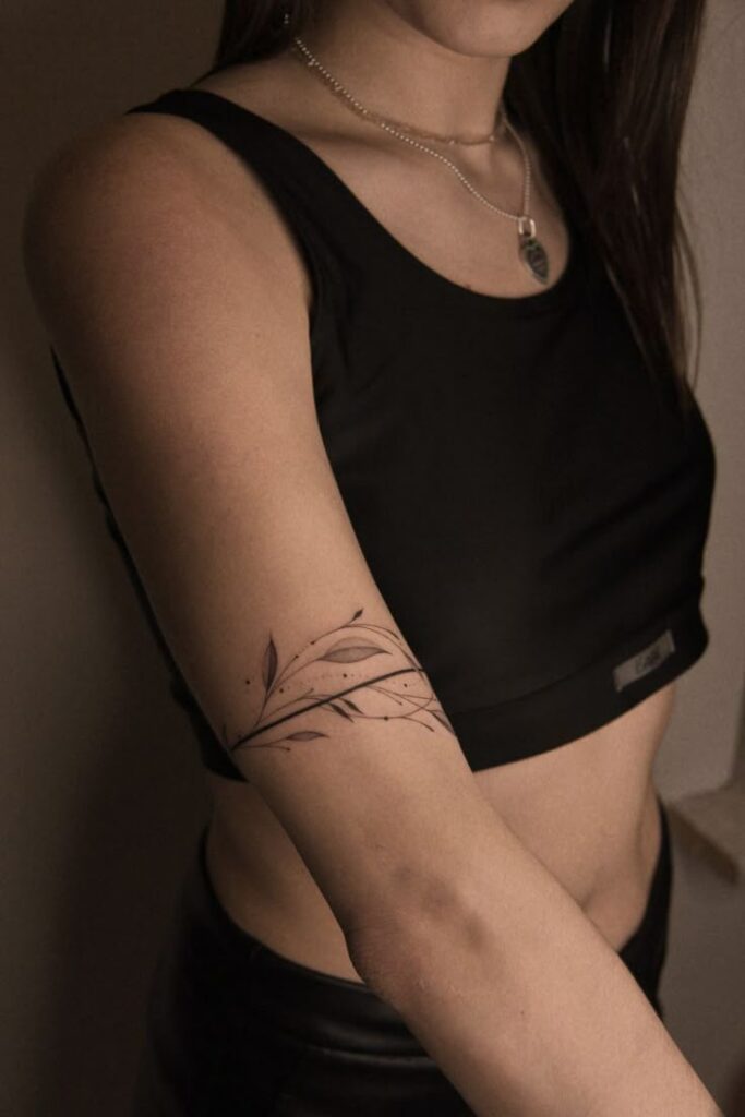Minimalist leaf tattoo on woman's upper arm, paired with a black tank top and silver necklace.