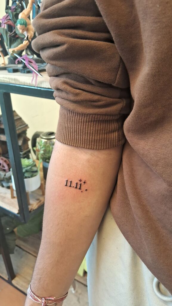 Minimalist 11.11 tattoo on forearm with small stars, brown sleeve above, indoor setting with plants in background.