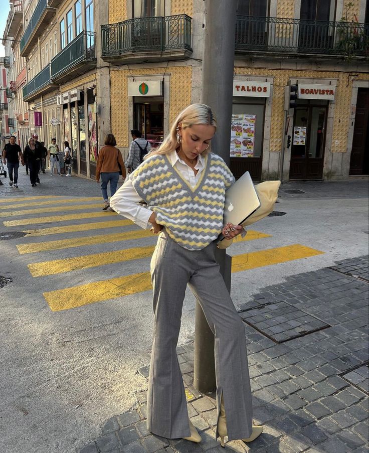 Fashionable woman in gray flared pants and patterned vest stands on city street with laptop, embracing urban style.