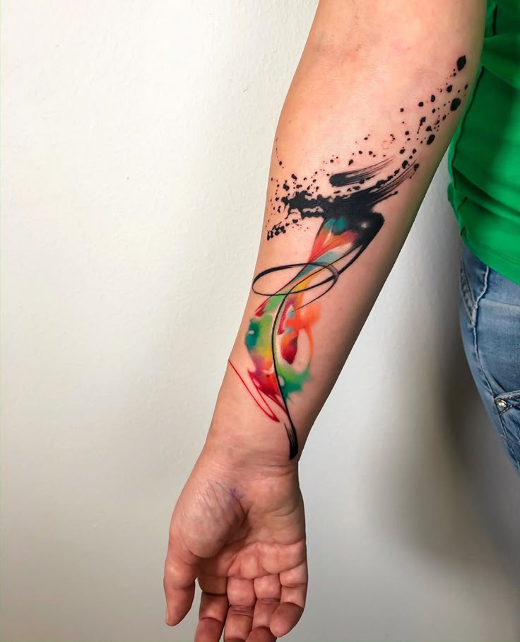 Abstract watercolor tattoo on forearm with black ink splatters and colorful swirls.