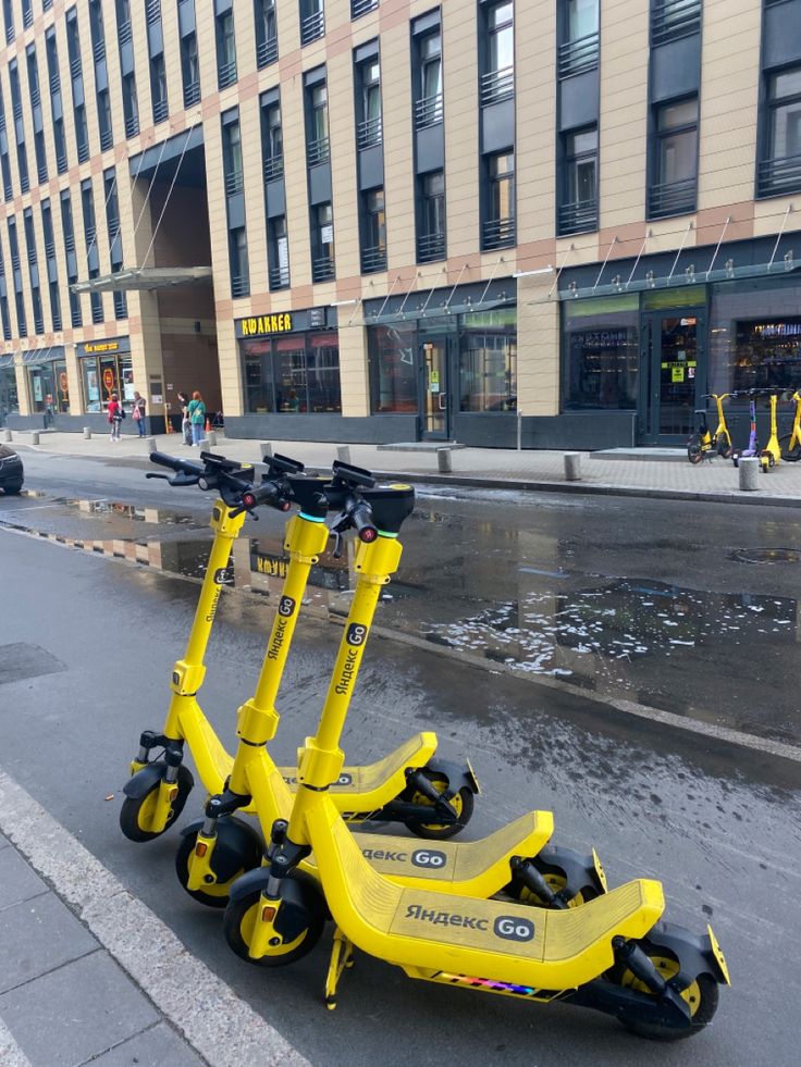 Three yellow rental scooters parked on a rainy urban sidewalk in front of a building.