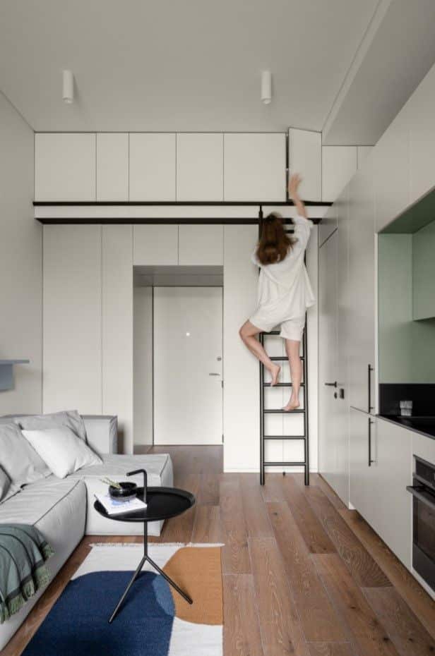 Woman reaches high cabinet on ladder in modern living room with sleek design and wooden flooring.