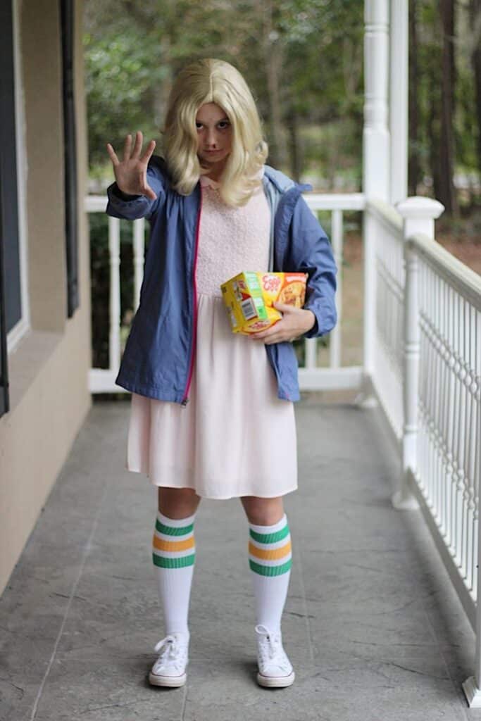 Person in costume with blonde wig and socks, holding waffles and snacks on a porch.