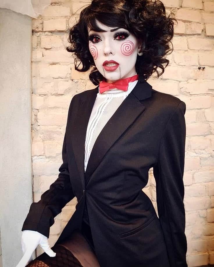 Person in curly wig and black suit with red bow, dramatic makeup featuring spiral cheek designs, standing against a brick wall.