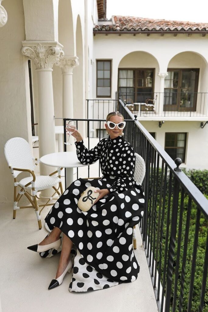 Woman in a polka dot dress enjoys a drink on a stylish balcony with white furniture and classic architecture.
