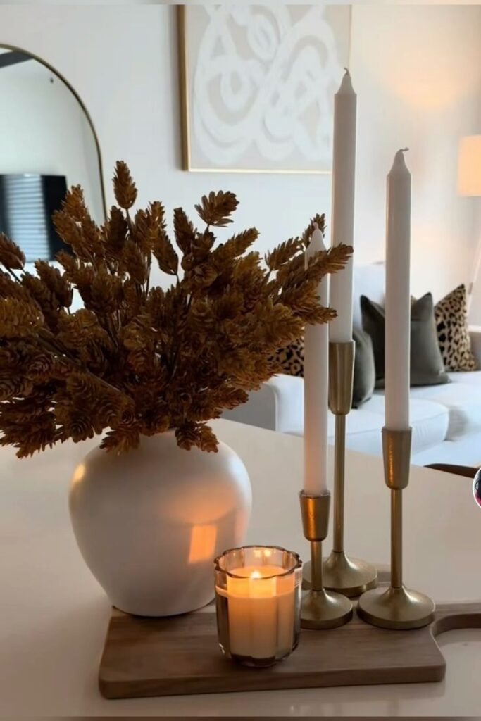 Cozy living room decor with white vase, dried flowers, white candles, and soft lit candle on a wooden tray.
