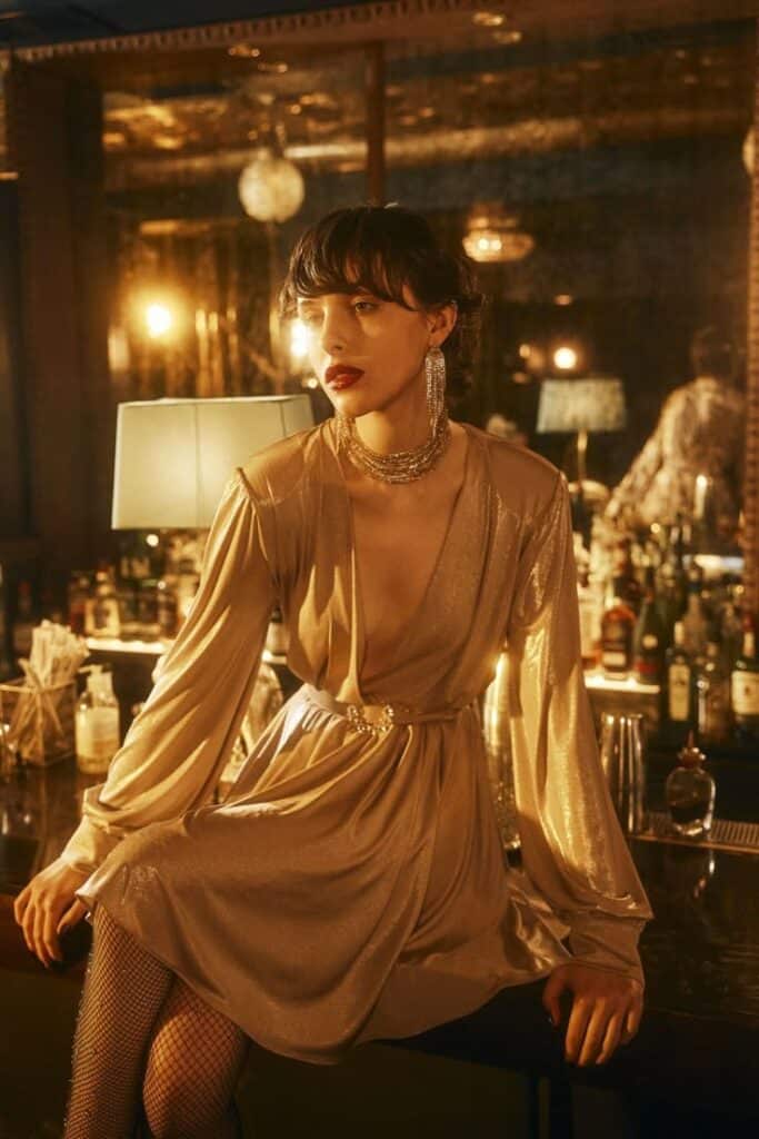 Person in elegant gold dress and statement jewelry sitting on bar counter in dim, stylish venue.